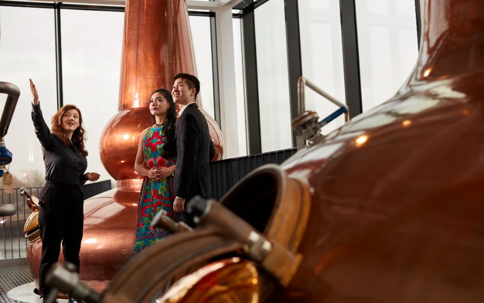 Three people standing inside a brewery or distillery with large copper brewing tanks.