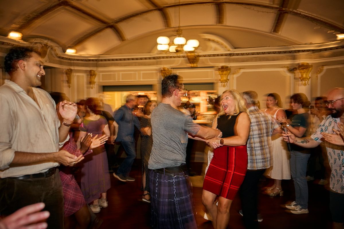 A man wearing a kilt and a woman in a red dress are dancing energetically.