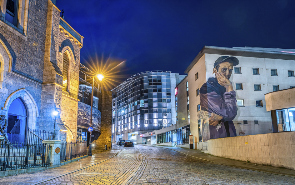 An illuminated cobbled street, curved glass building, and a large mural.