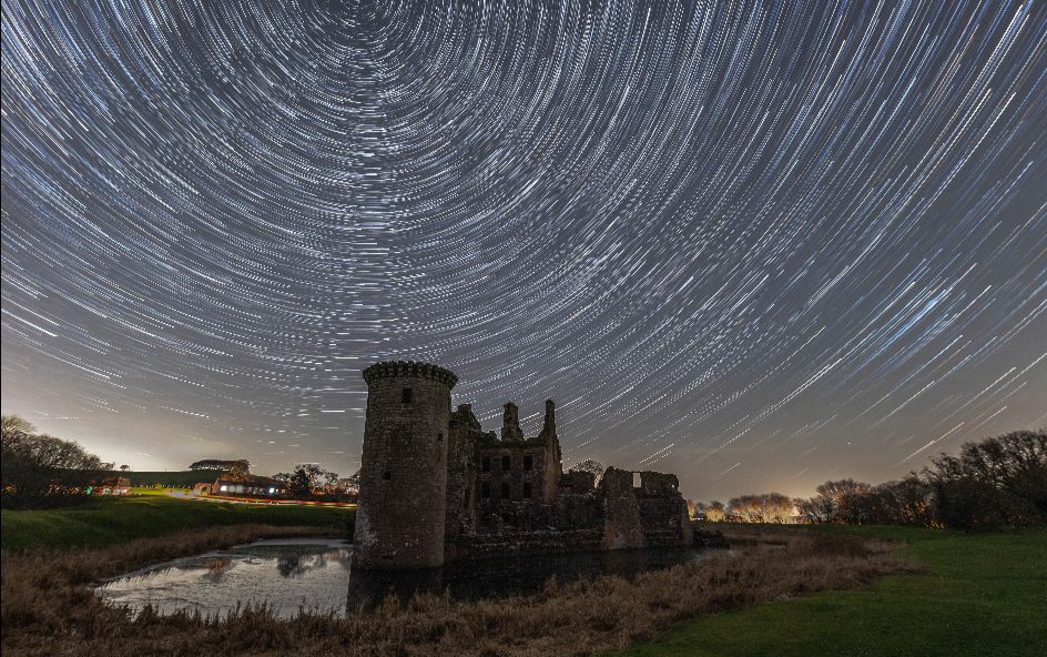 A historic castle ruin under a night sky with star trails forming circular patterns around a central point.