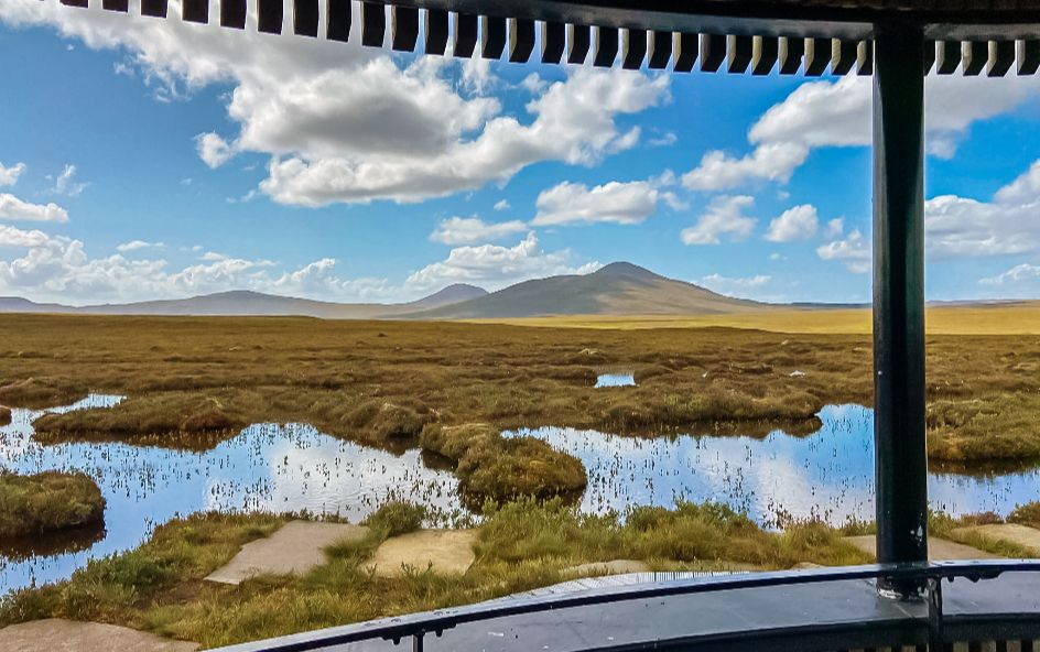 A vast marshland with patches of water reflecting blue sky and scattered clouds, framed by a curved railing and striped roof edge.