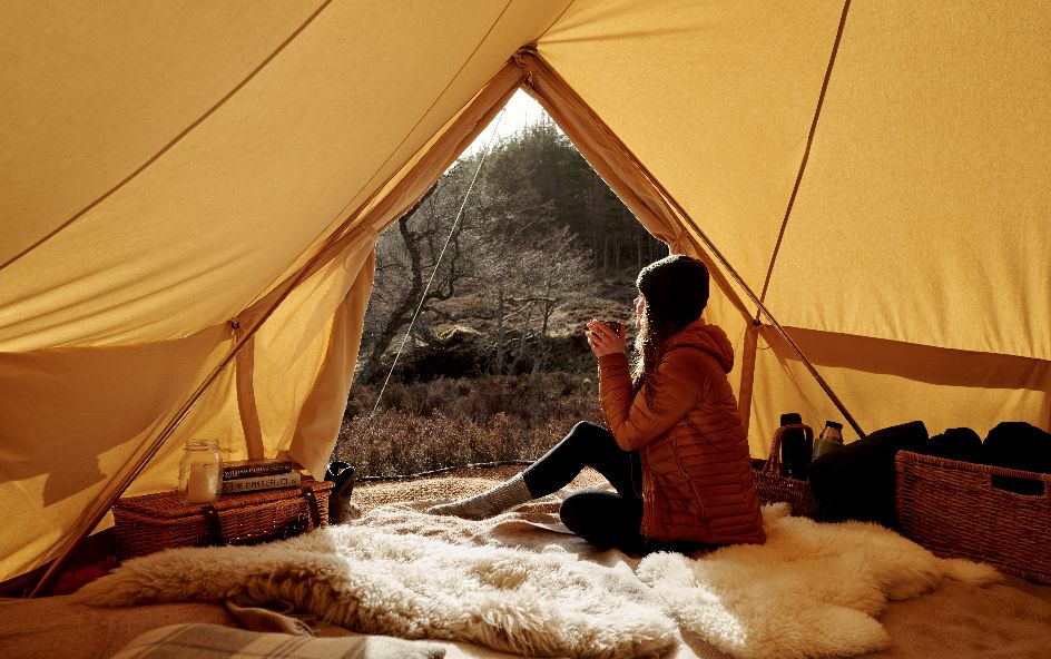 A person sitting inside a tent, facing an outdoor natural landscape with trees and shrubs.
