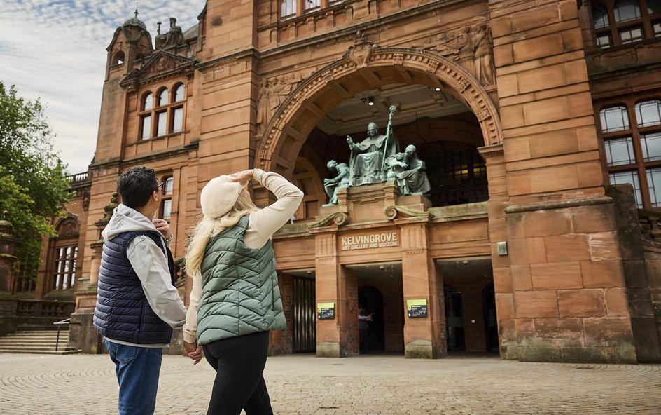 Two people standing outside a large, historic red-brick building with an arched entrance labeled