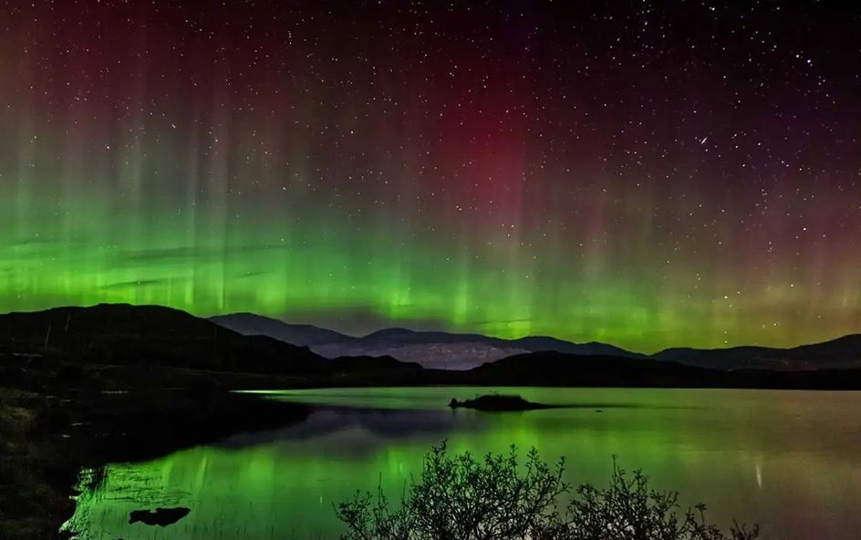 Green, purple and yellow Northern light skies shimmering above a loch and mountains.
