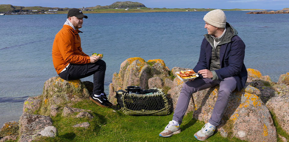 two people sit on rocks on the coastline with food