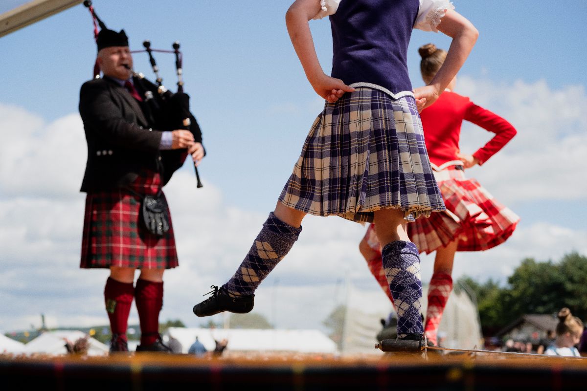 Traditional Scottish dancers performing on an outdoor stage with a bagpiper playing in the background.