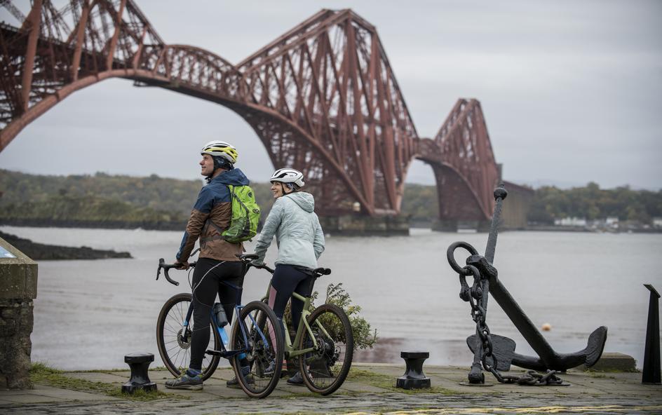 Two cyclists near water, with a steel bridge in the background.