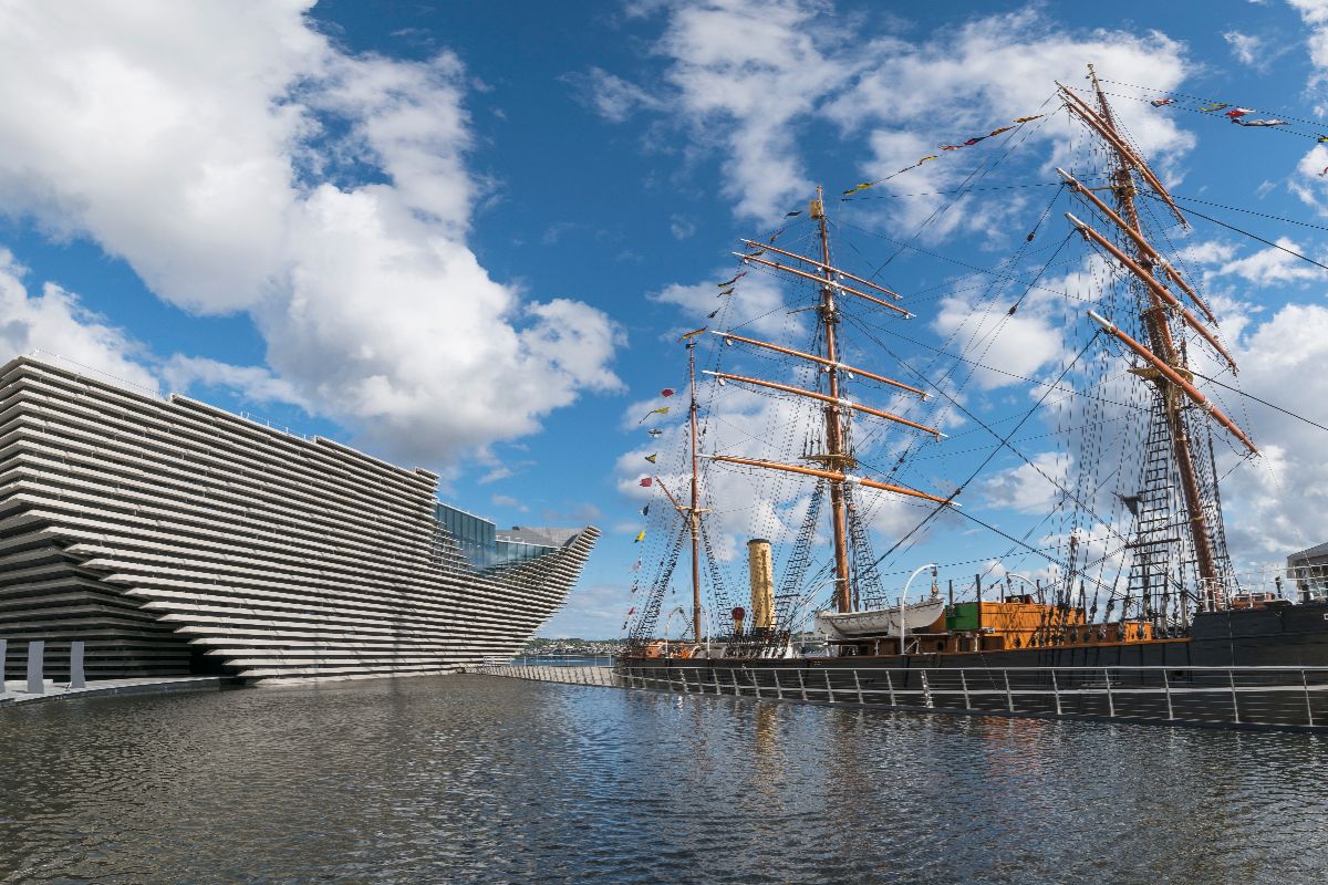 A modern, angular museum building next to a historic tall ship and a body of calm water.