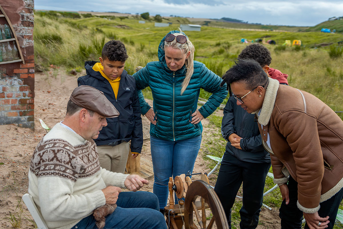 four people look on as a weaver spins wool on a wheel