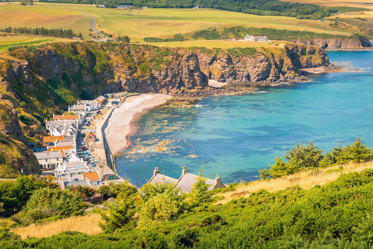 A coastal village nestled at the base of rugged cliffs with clear turquoise waters along a curved shoreline.