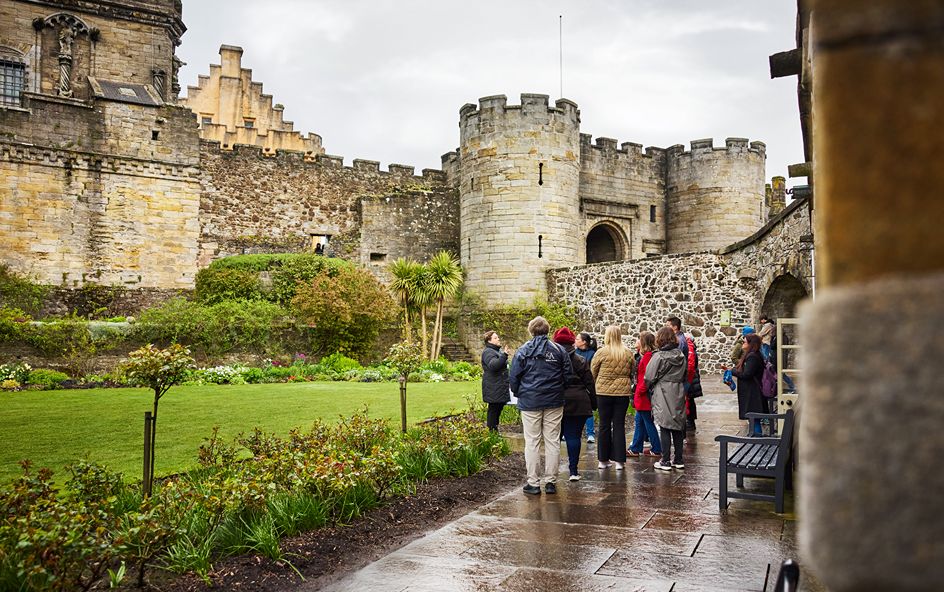 A group of tourists gathered near a historic stone castle. with battlements and a round tower.