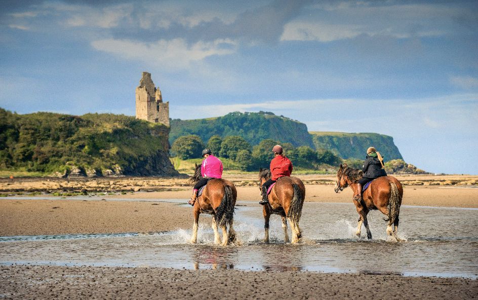 Three horses galloping on beach coast with castle ruin in back.