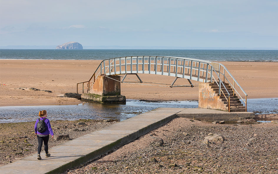 A person walking towards a bridge over some water, on a beach