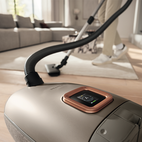 Close up on the LCD screen feature of the Miele Guard L1 with a background of a man vacuuming his living room.