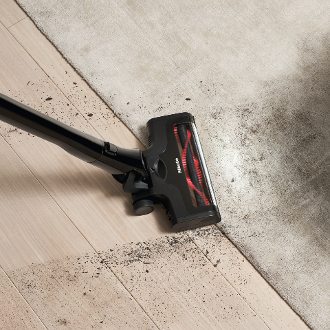 Vacuum cleaner head removing dirt from wood floor and light rug beside a gray sofa, showing cleaning action.