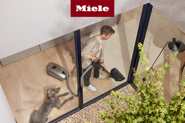 Man seen through a window vacuuming hardwood flooring using the Miele Guard L1 with dog laying next to him 