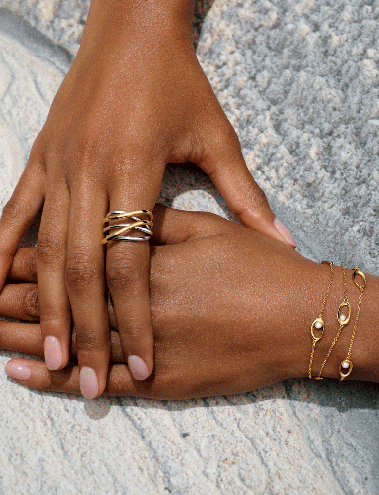 A pair of hands overlap and display an intertwined ring and pearl bracelet.